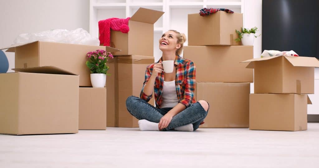 people, moving new place and repair concept happy beautiful young woman with many cardboard boxes sitting on floor with Cup at home